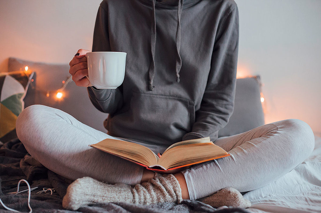 Frau sitzt auf Bett im Schneidersitz, liest Buch und trinkt Kaffee, Wohlfühlatmosphäre mit Holz in einem KAMPA Fertighaus 